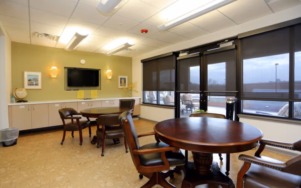 A well-lit interior room with round wooden tables and leather chairs on wheels. The room has a green accent wall with a mounted flat-screen TV, two wall sconces, framed pictures, and a long cabinet underneath. Large windows with dark roller shades cover one wall, and a door leads to an outdoor patio area with seating.