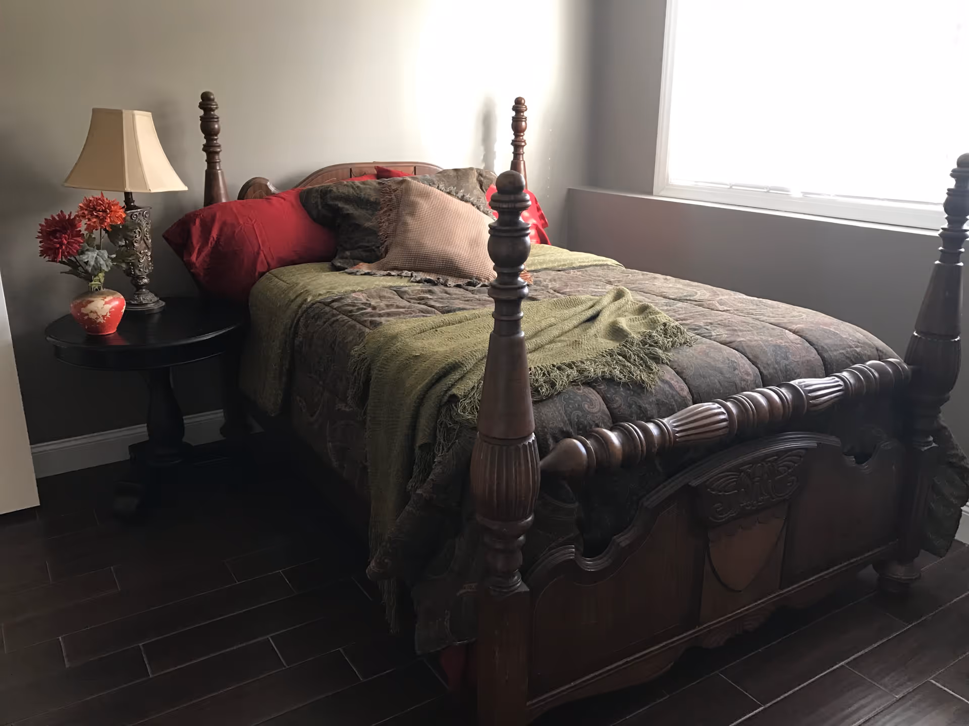A bedroom with a wooden four-poster bed covered with a patterned comforter, green throw blanket, and multiple pillows. Next to the bed is a round black nightstand with a decorative lamp and a vase with red flowers. A large window with white blinds is on the right side, letting in natural light.