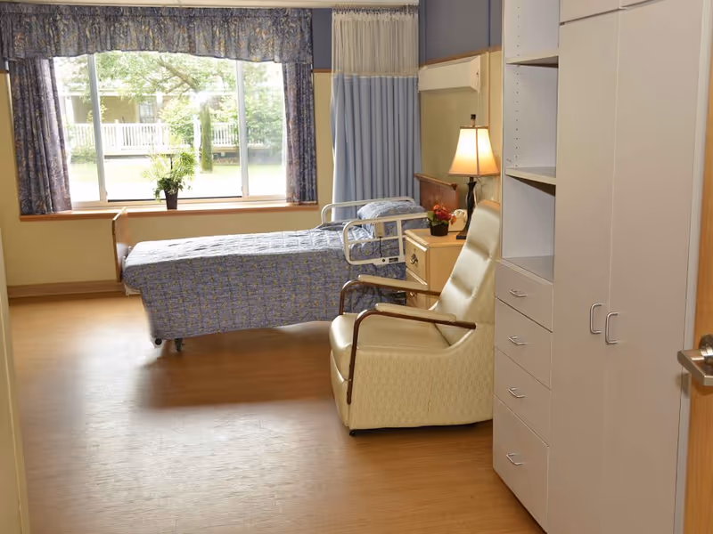 A bright and clean rehabilitation center room with a single hospital bed covered in a blue patterned blanket, a beige recliner chair, a wooden nightstand with a lamp and a small plant, and a large window with blue floral curtains letting in natural light. There is also a white storage cabinet with drawers and shelves on the right side of the room.