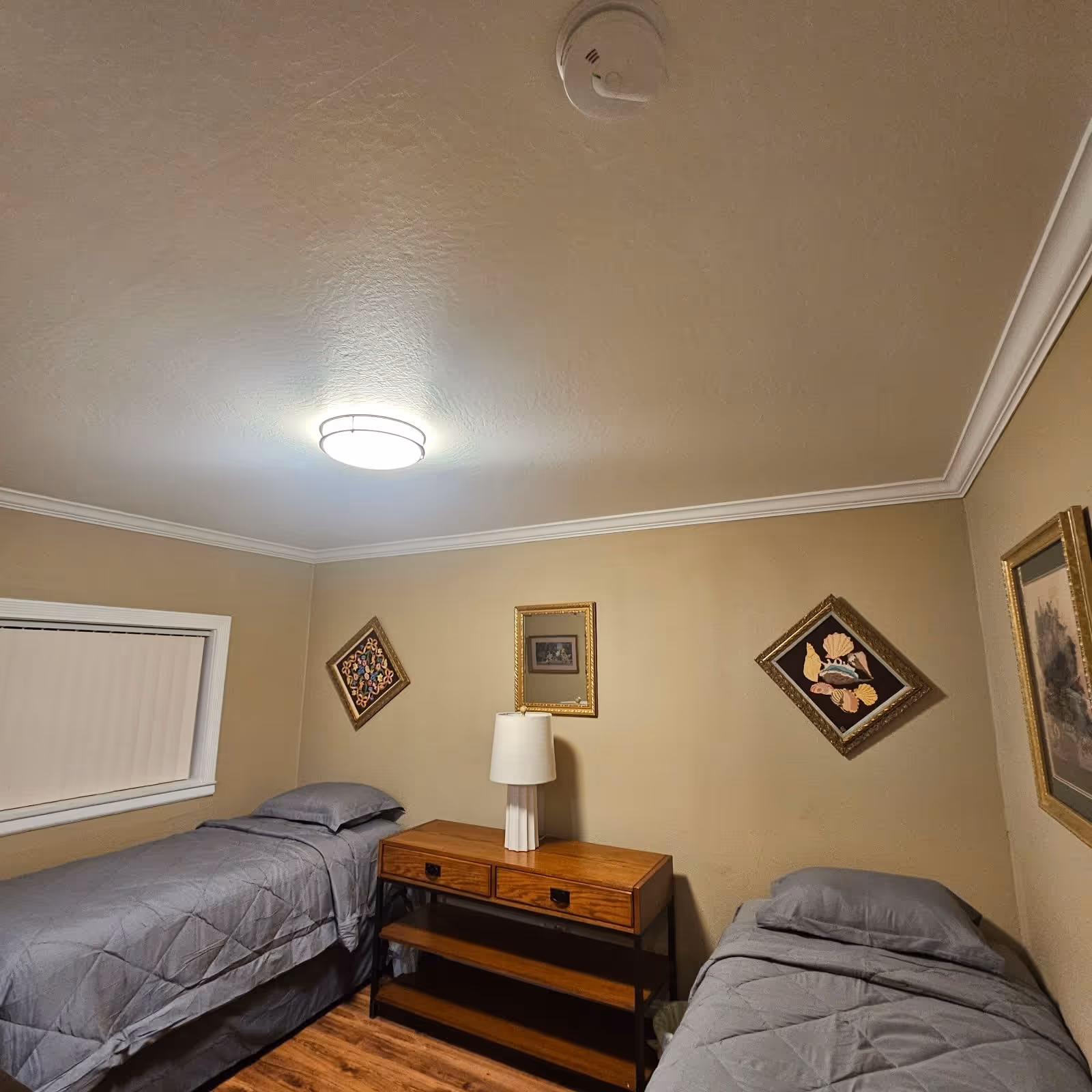 Two twin beds with gray bedding flank a wooden nightstand and lamp in a small bedroom decorated with framed wall art and a ceiling light.