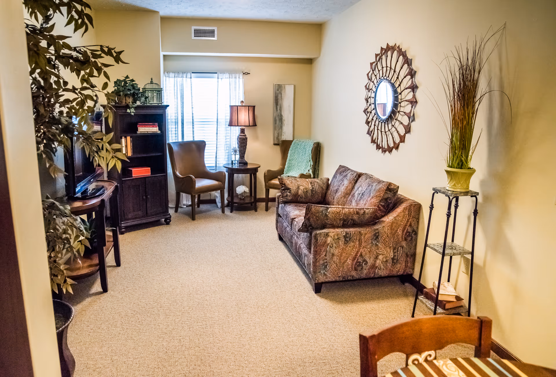 A cozy living room with a patterned sofa, two brown armchairs, a small round table with a lamp, a dark wooden cabinet with books and decorative items, a large leafy plant, and a decorative mirror on the wall. The room has beige walls and carpeted floor with a window covered by sheer curtains.