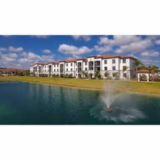 White multi-story building with red tile roofs overlooking a pond with a central fountain under a partly cloudy sky.