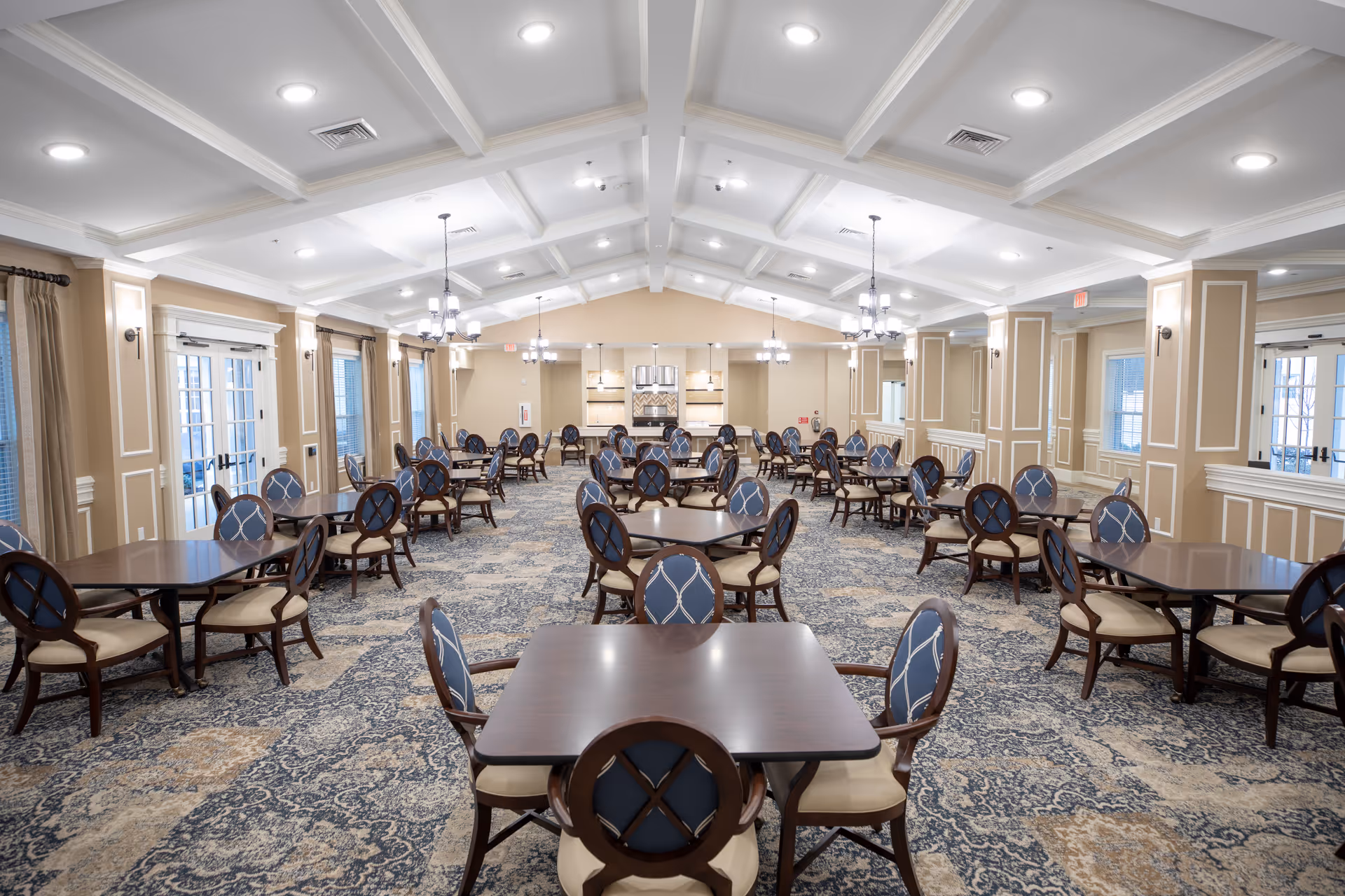 A spacious dining room with multiple dark wooden tables and blue cushioned chairs arranged neatly. The room features a patterned carpet, beige walls with white trim, large windows with curtains, and several chandeliers hanging from a white coffered ceiling. There are double glass doors on both sides of the room.