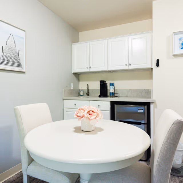 Small dining area with a round white table and two upholstered chairs. On the table is a small white vase with pink flowers. Behind the table is a kitchenette with white cabinets, a small sink, a coffee maker, and a mini refrigerator. The walls are light-colored with framed artwork hanging on them.