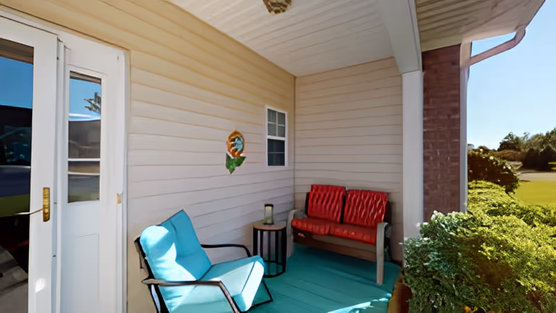 A covered outdoor porch area with a light blue cushioned chair, a small round side table, and a red cushioned bench. The porch has beige siding, a small window, and overlooks a green lawn with bushes.