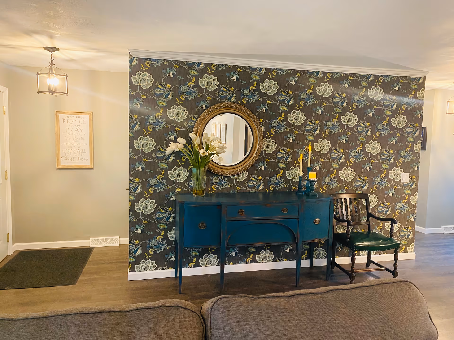 A cozy interior space featuring a floral patterned accent wall with a round mirror hanging above a teal sideboard. On the sideboard, there is a vase with white flowers and two yellow candles. To the right of the sideboard is a wooden chair with a green cushion. The floor is wood, and a gray couch is partially visible in the foreground. A framed inspirational quote hangs on a plain wall to the left, near a door and a ceiling light fixture.