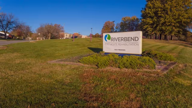 Outdoor view of a grassy area with a sign that reads 'Riverbend Post Acute Rehabilitation 7850 Freeman' surrounded by bushes, with trees and a clear blue sky in the background.