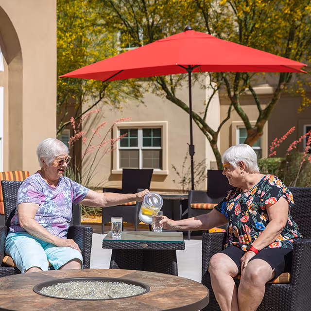 Two elderly women sitting outdoors on wicker chairs under a red patio umbrella. One woman is pouring water with lemon slices from a pitcher into a glass held by the other woman. They are smiling and enjoying a sunny day in a courtyard with trees and a beige building in the background.