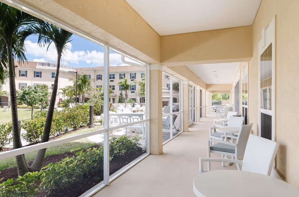 A covered outdoor patio area with white chairs and small round tables along the right side, overlooking a landscaped garden with palm trees and bushes. In the background, there is a multi-story beige building with windows and a red-tiled roof under a blue sky with some clouds.