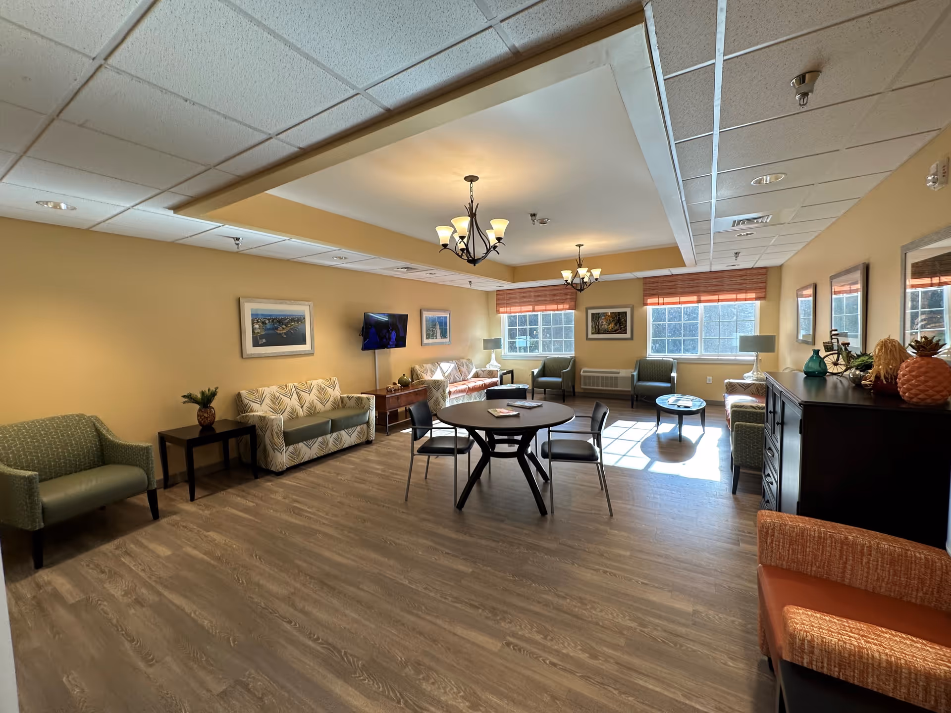 Communal resident lounge with sofas, armchairs, a round table, TV, and large windows.