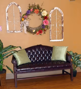 A dark brown tufted leather loveseat with two green textured pillows is placed against a beige wall. Above the loveseat, there is a decorative floral wreath with colorful flowers and two white distressed wooden wall hangings with scrollwork designs. Two green potted plants are positioned on either side of the loveseat.