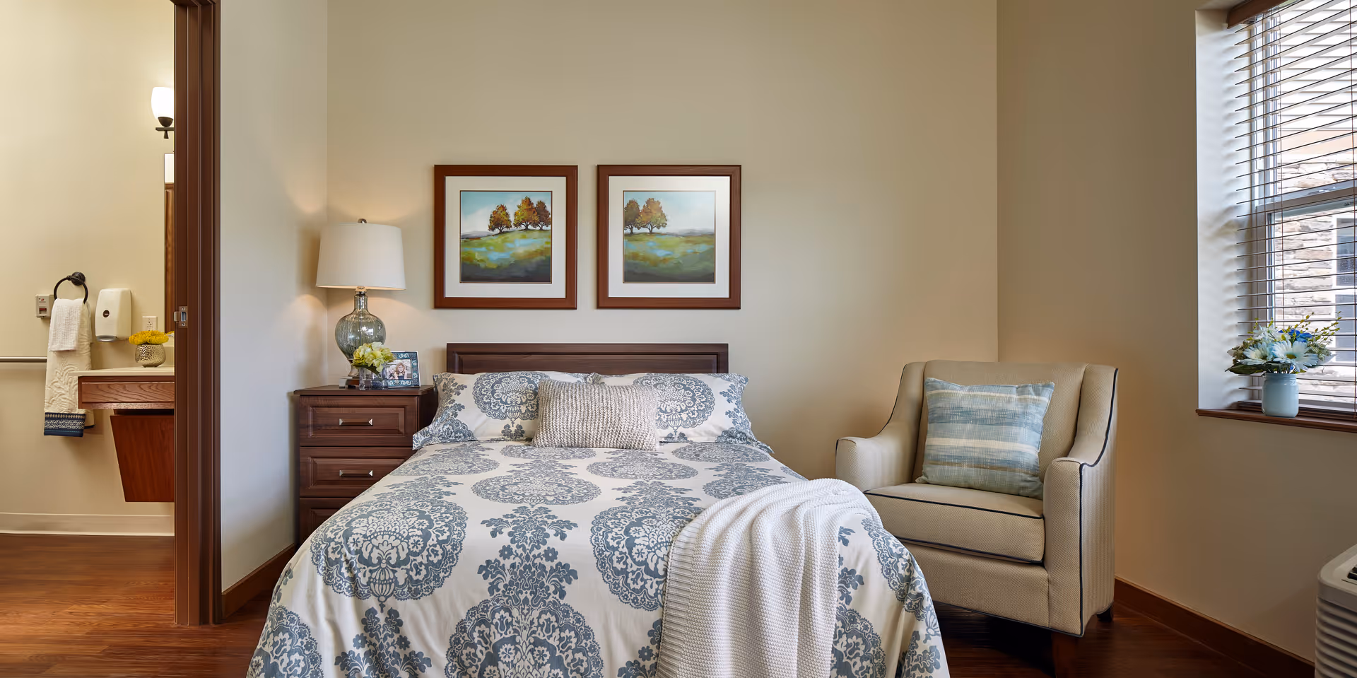 A cozy bedroom with a bed covered in a white and blue patterned comforter, a beige armchair with a blue striped pillow, a wooden nightstand with a lamp and framed photos, two landscape paintings on the wall above the bed, and a window with blinds letting in natural light. A bathroom is partially visible through an open door on the left side.