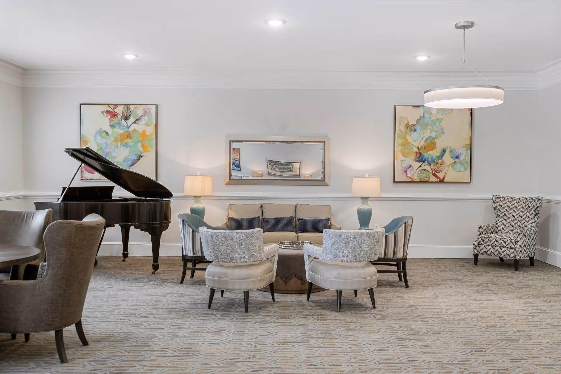 A spacious and well-lit living room featuring a grand piano on the left, a beige sofa with blue and gray cushions in the center, two light-colored armchairs with patterned upholstery, and two blue armchairs. The room has two table lamps on either side of the sofa, a large rectangular mirror above the sofa, colorful abstract paintings on the walls, and a modern circular ceiling light fixture. The floor is carpeted with a subtle pattern.