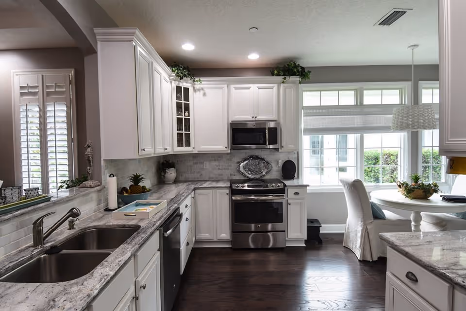 A modern kitchen with white cabinets, marble countertops, and stainless steel appliances including a stove and microwave. There is a double sink on the left side and a small round dining table with two upholstered chairs near large windows letting in natural light. The floor is dark hardwood and there are decorative plants on top of the cabinets and table.