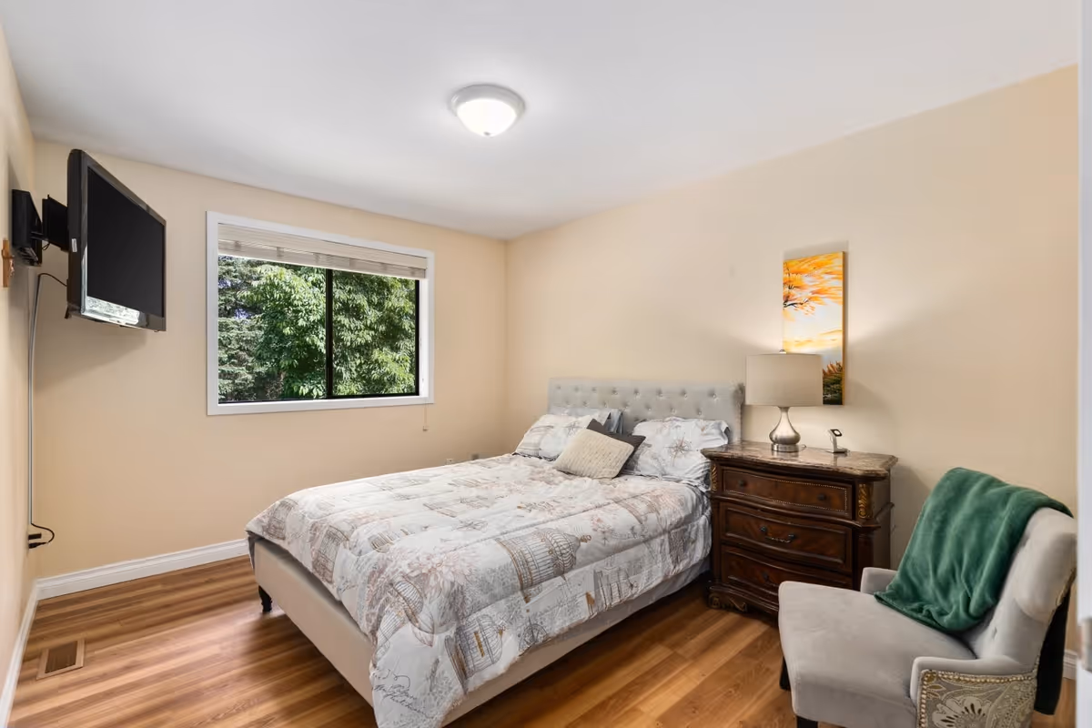 A cozy bedroom with a bed covered in a patterned comforter and multiple pillows. There is a wooden nightstand with a lamp and a small decorative item next to the bed. A cushioned chair with a green throw blanket is positioned near the nightstand. A window with a view of green trees is on one wall, and a flat-screen TV is mounted on the opposite wall. The room has light-colored walls and wood flooring.