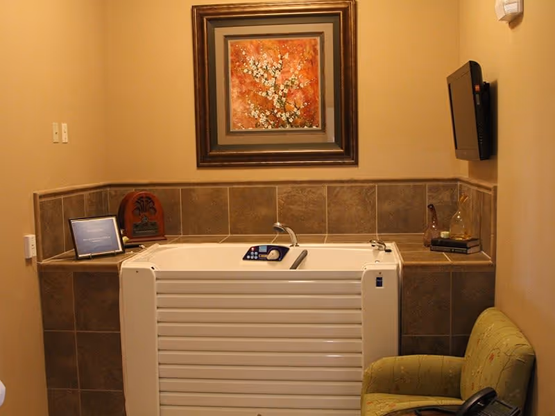 A small bathtub with a control panel and faucet, surrounded by brown tiled walls. Above the tub is a framed painting of white flowers on an orange background. To the right of the tub is a small shelf with decorative items and a wall-mounted TV. A green upholstered chair is partially visible in the lower right corner.