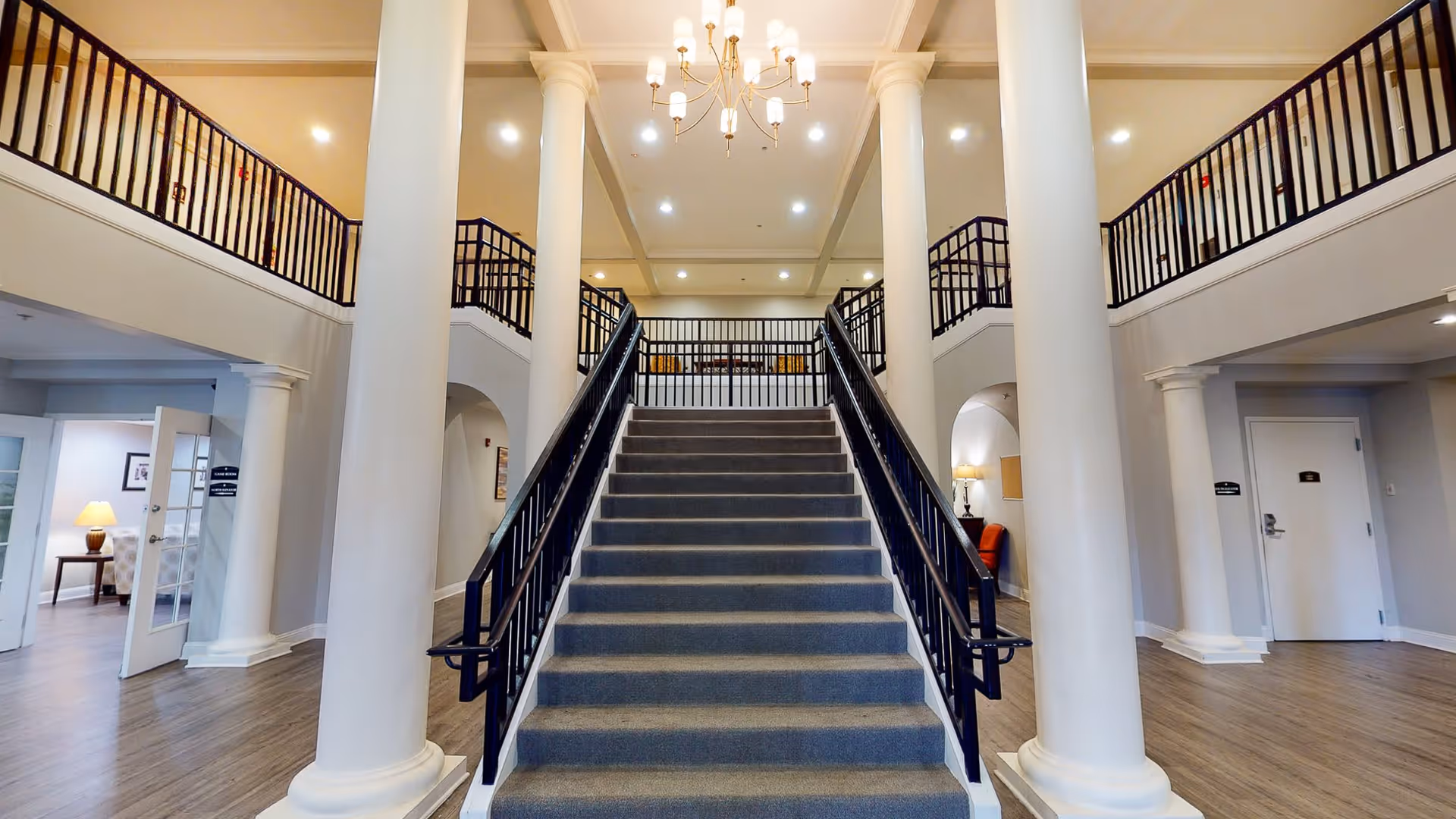 Interior view of a senior living facility featuring a wide staircase with black railings leading to an upper floor. The space has tall white columns, wooden flooring, and a chandelier hanging from the ceiling. There are open doorways on either side of the staircase, with some furniture and lamps visible inside the rooms.