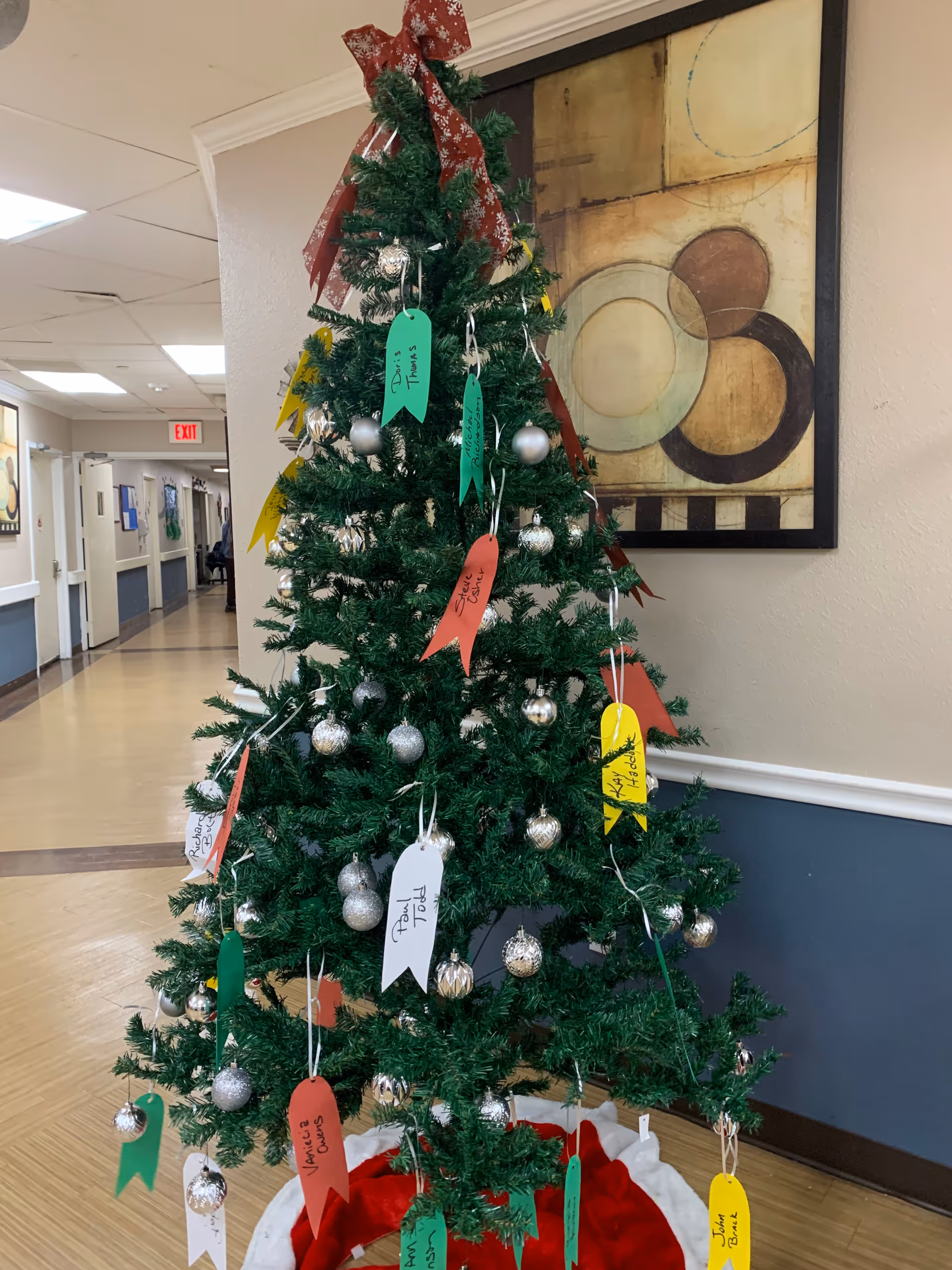 A decorated Christmas tree with silver ornaments and colorful paper tags hanging from its branches, placed in a hallway of a nursing center. The tree has a red bow on top and a red and white tree skirt at the base. The hallway has beige walls with blue wainscoting, a framed abstract painting on the wall behind the tree, and an exit sign visible in the distance.