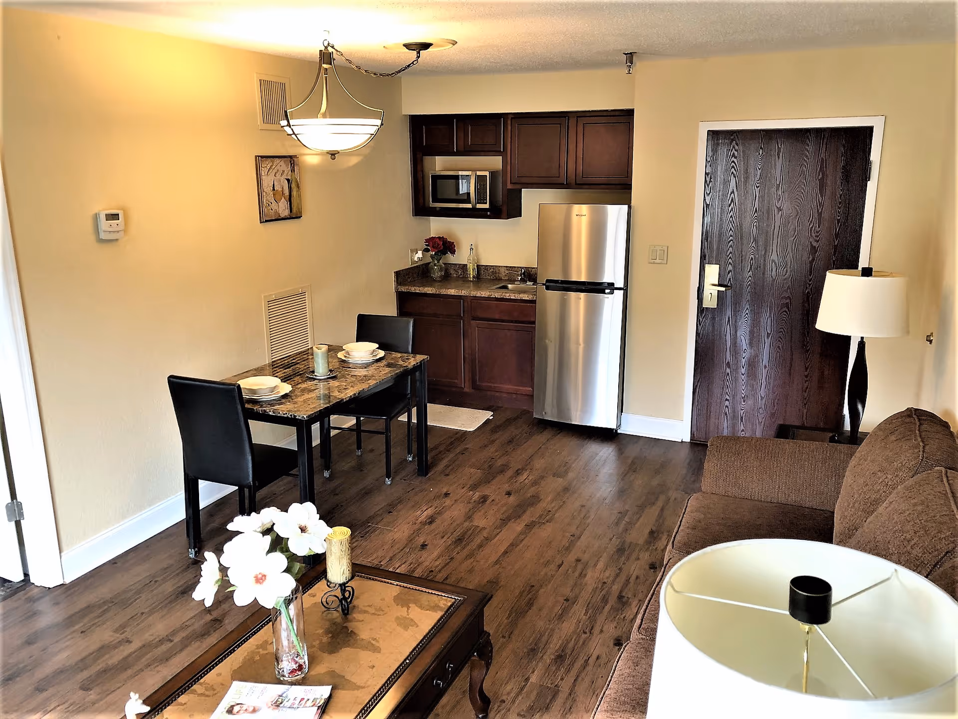 Interior view of a senior community apartment showing a small kitchen area with dark wood cabinets, a stainless steel refrigerator, and a microwave. Adjacent to the kitchen is a dining table set for two with black chairs. The room has wood flooring, a brown sofa, a coffee table with a vase of white flowers, and a lamp. A dark wooden door is visible in the background.