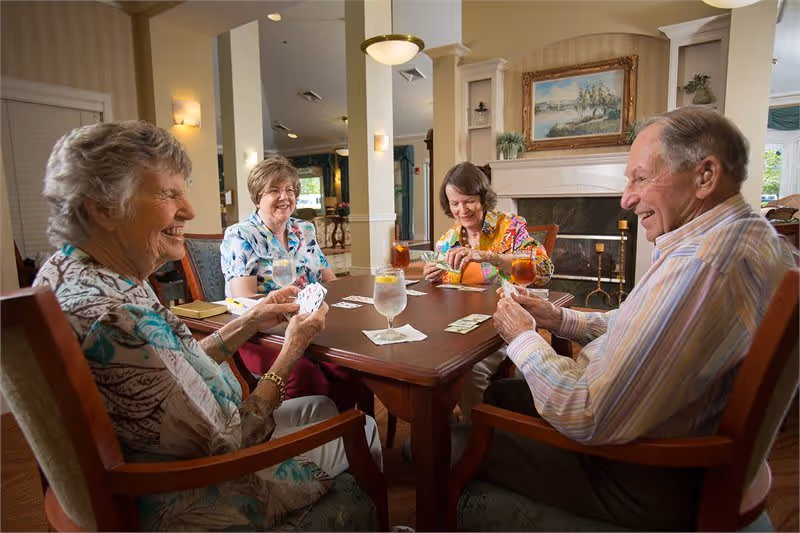 Four elderly people sitting around a wooden table playing cards in a well-lit room with a fireplace and framed painting in the background. They appear to be enjoying themselves, with drinks on the table including water and iced tea.