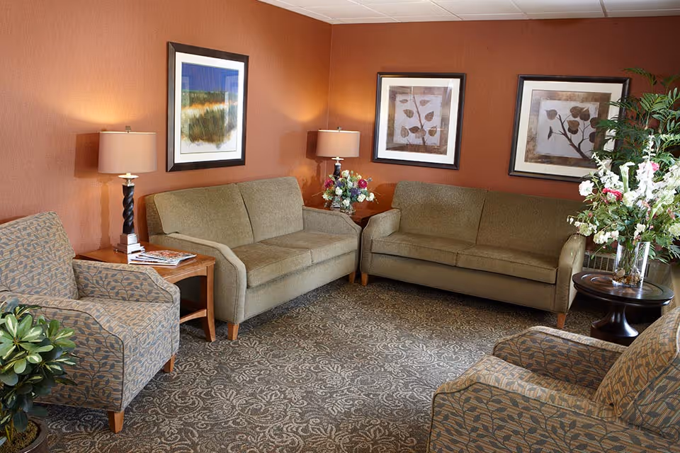 A cozy living room with two beige sofas, two patterned armchairs, a wooden side table with a lamp and magazines, floral arrangements on side tables, and three framed botanical prints on the orange walls.