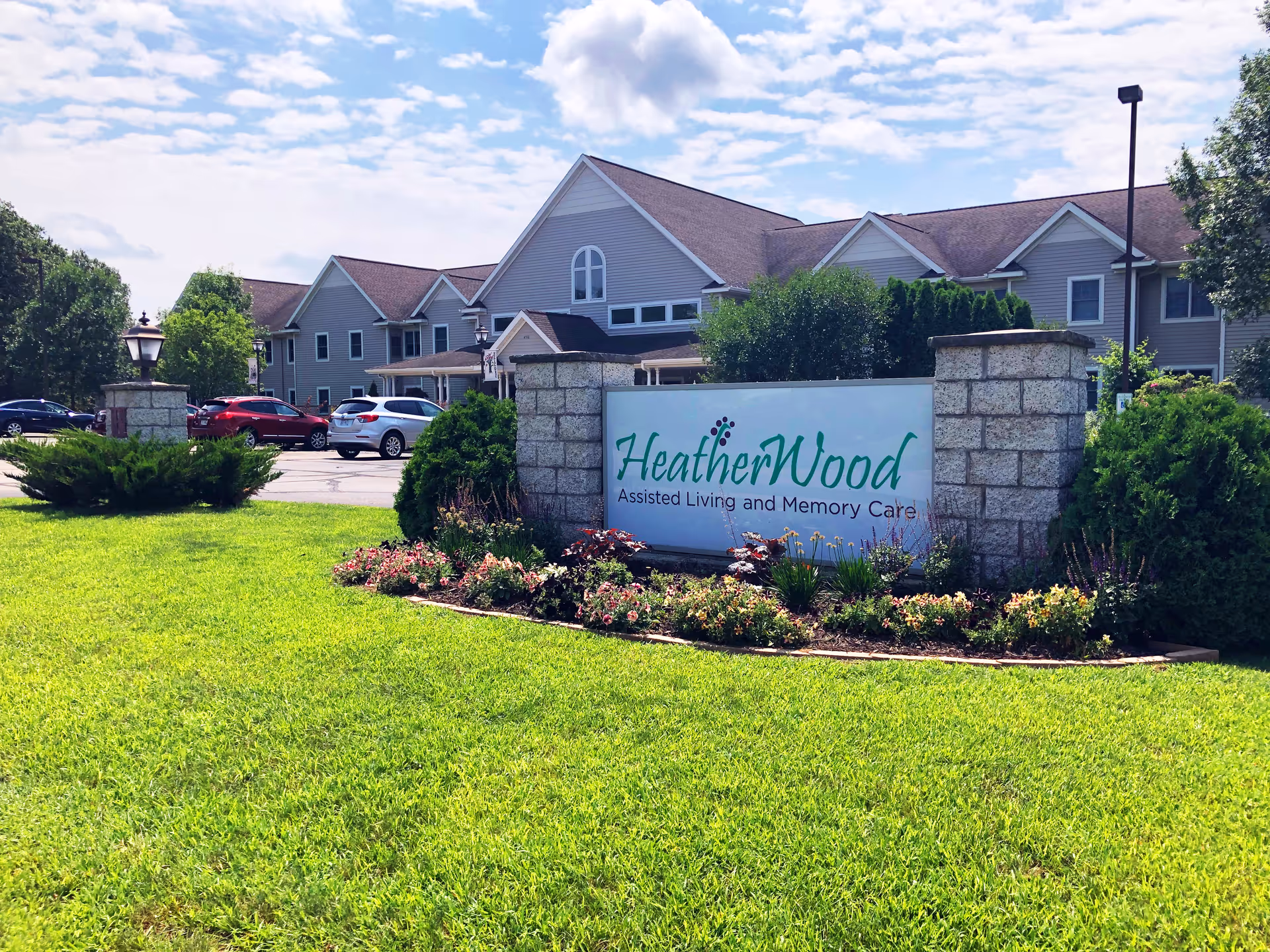 Exterior view of HeatherWood Assisted Living and Memory Care facility with a large sign in front surrounded by flowers and greenery. The building is a two-story structure with gray siding and multiple windows. Several cars are parked in the parking lot behind the sign under a partly cloudy sky.