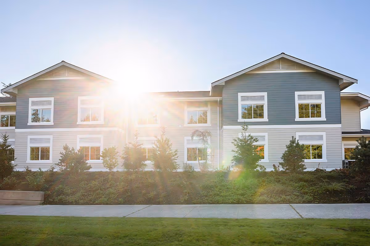 Exterior view of a two-story building with blue and white siding, multiple windows, and landscaping with bushes and small trees in front. The sun is shining brightly behind the building, creating a lens flare effect.