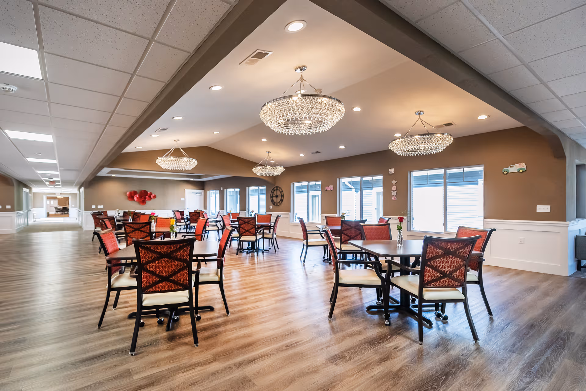 Spacious dining room with multiple tables and red-upholstered chairs, crystal chandeliers, and wood-look flooring.