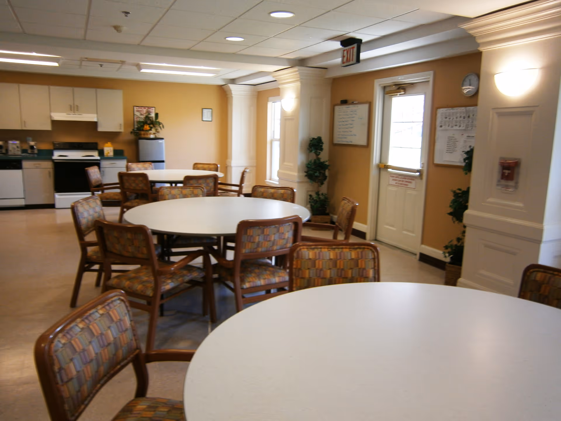 A senior living community dining area with round tables and cushioned chairs arranged around them. The room has a kitchen area with white cabinets, a stove, a refrigerator, and a dishwasher. The walls are painted a warm beige color, and there are plants and wall decorations. An exit door is visible on the right side of the room.