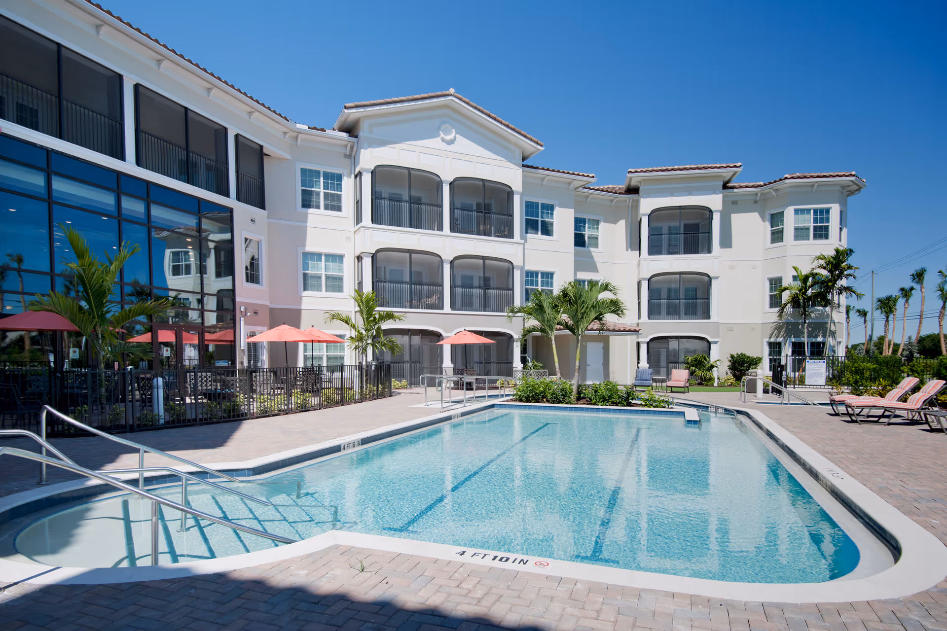 Outdoor swimming pool area at Allegro Senior Living in Parkland, FL, featuring a clear blue pool with steps and handrails, surrounded by a paved deck with lounge chairs and umbrellas. The background shows a three-story white building with balconies and large windows under a clear blue sky.