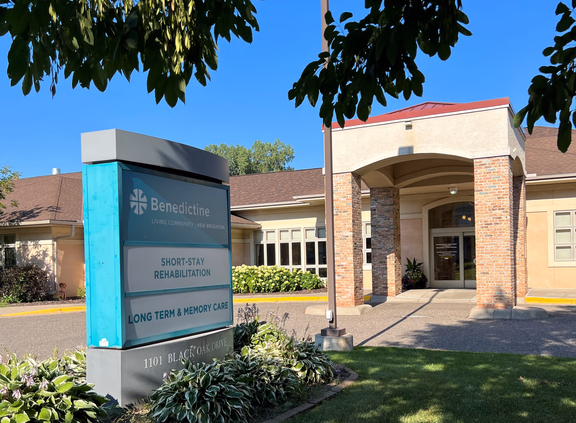 Front entrance of Benedictine Living Community - New Brighton with a teal sign listing short-stay rehabilitation and long-term & memory care.