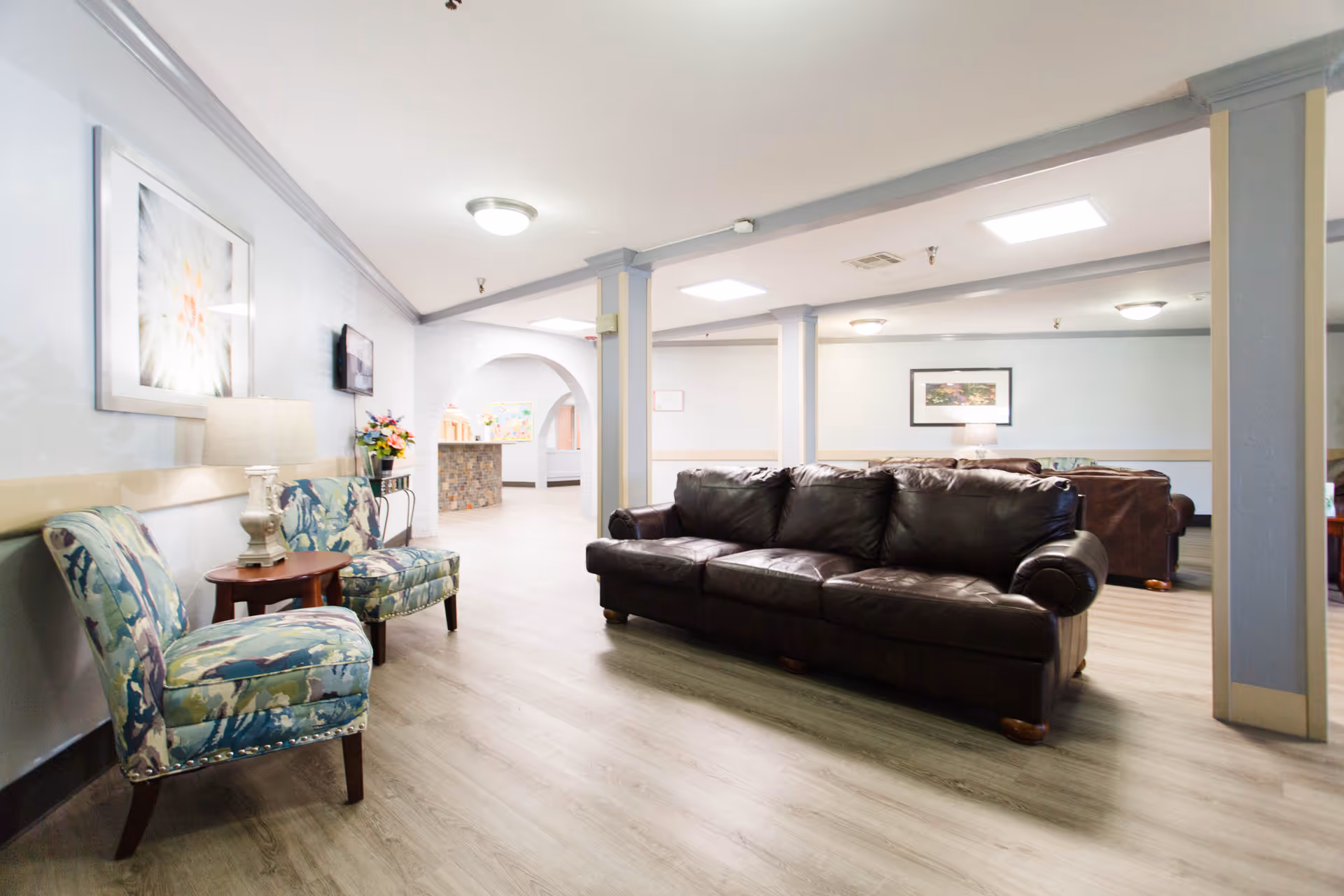 A spacious and well-lit living room area in a senior living facility featuring a dark brown leather sofa, two floral patterned armchairs with a small wooden side table and lamp between them. The room has light-colored wood flooring, white walls with gray trim, framed artwork, and ceiling lights. In the background, there is an open archway leading to another area with a stone counter and more seating.