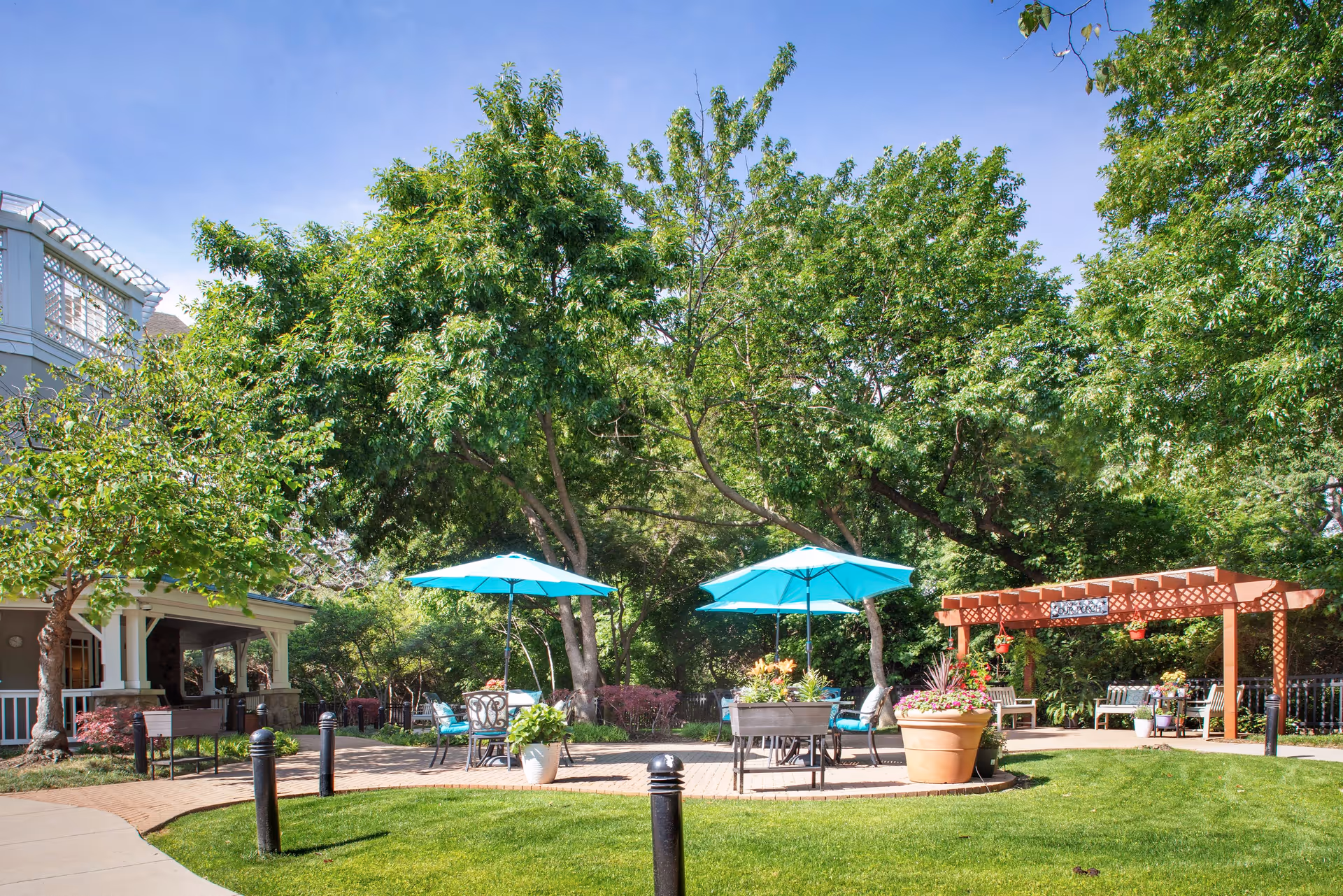 Outdoor garden area at Sunrise at Hillcrest with green grass, large trees, two blue patio umbrellas over tables with chairs, a wooden pergola with hanging plants, and a building porch visible on the left side.