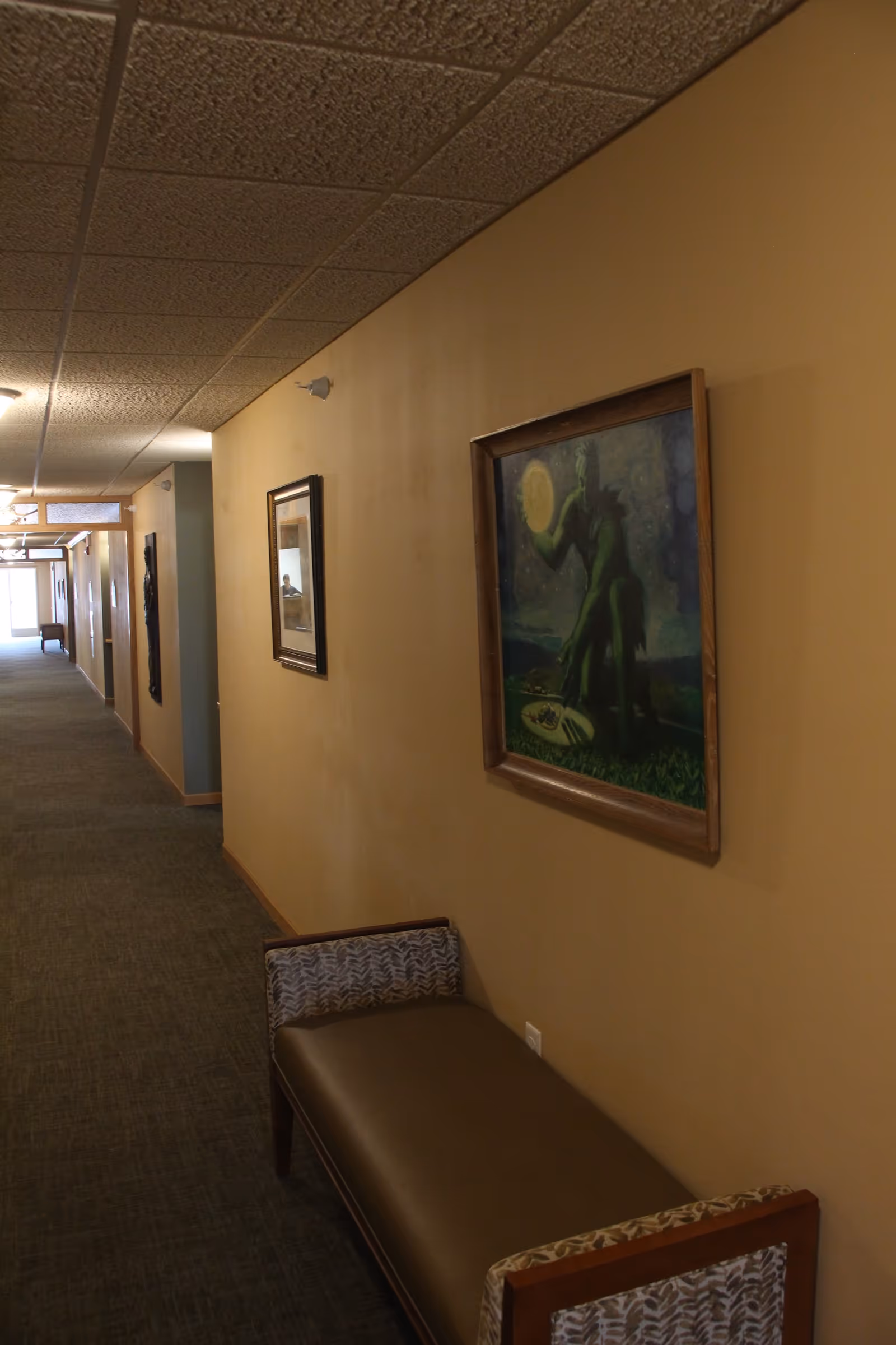 A long hallway with beige walls and a carpeted floor. On the right wall, there are framed paintings and a bench with patterned armrests. The ceiling has a grid pattern with recessed lighting. The hallway extends into the distance with doors and more artwork visible.