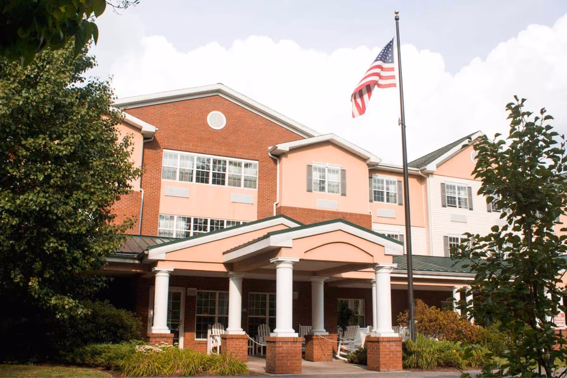 Exterior view of a senior living facility building with a covered entrance supported by white columns. The building has a combination of red brick and light pink siding with multiple windows. An American flag is flying on a flagpole in front of the entrance, and there are trees and shrubs around the building.