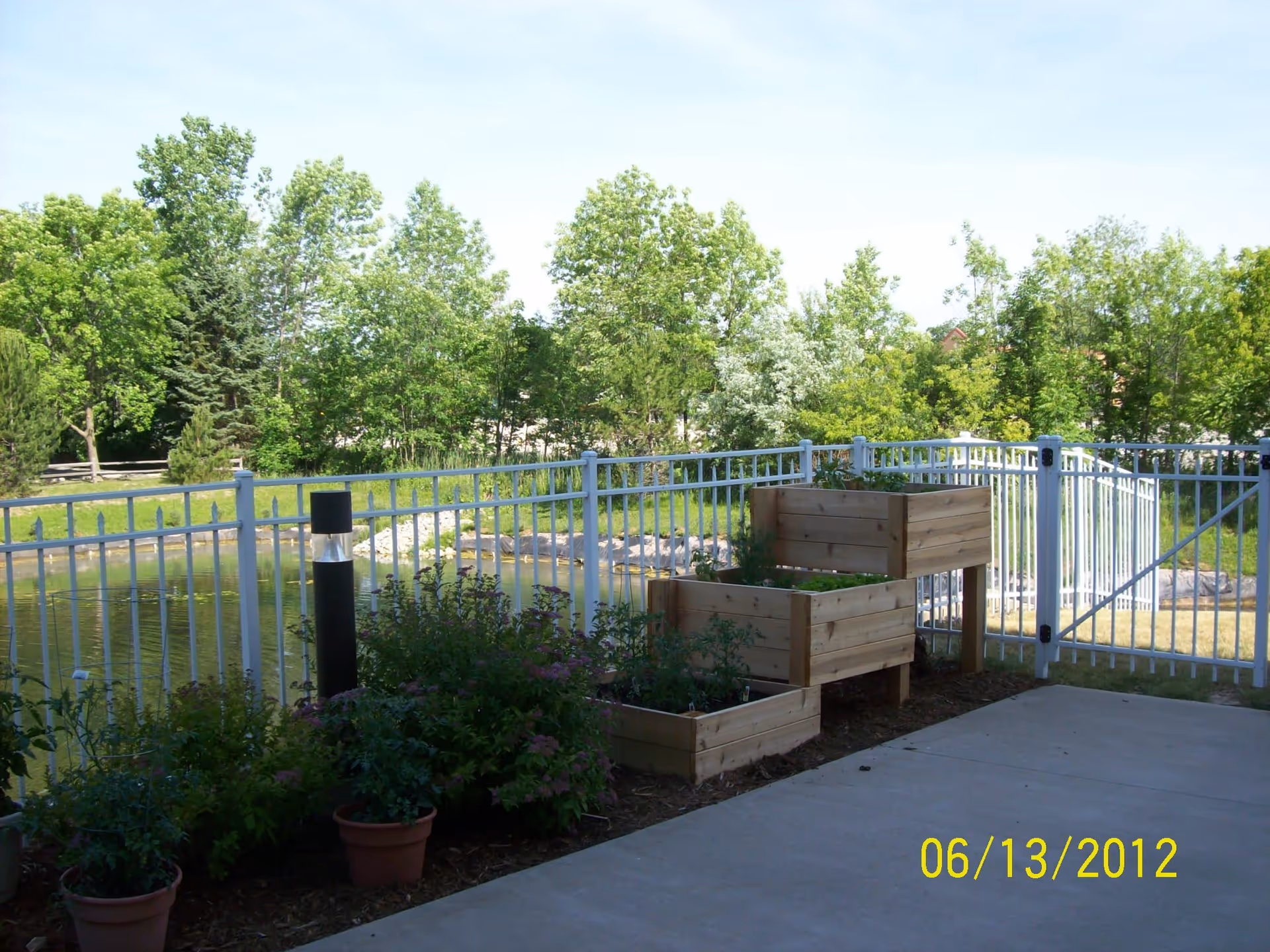 Outdoor patio with raised wooden planters beside a white metal fence overlooking a pond and trees.