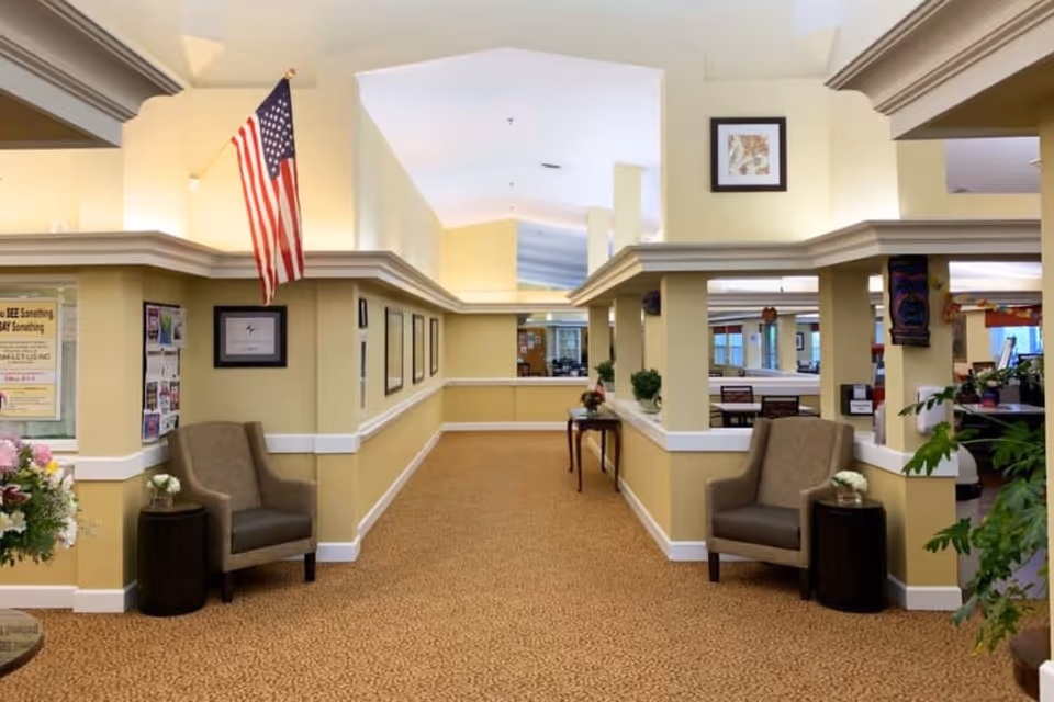Interior hallway of Lassen House Senior Living with beige walls and carpet. Two armchairs with small tables and flowers are placed on either side of the hallway. An American flag is mounted on the left wall. The hallway leads to a common area with tables and chairs visible in the background. Various framed pictures and plants decorate the space.