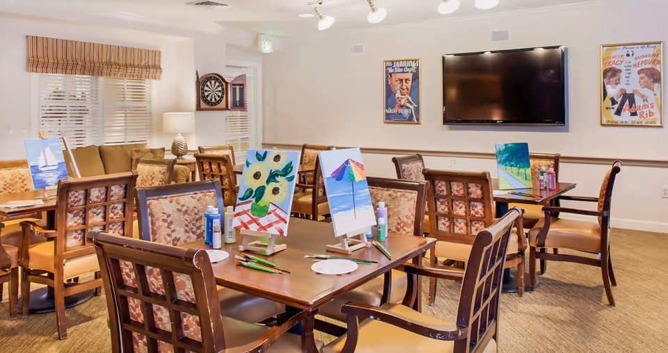 A well-lit activity room with several wooden tables and cushioned chairs arranged around them. Each table has small easels holding colorful paintings, paint bottles, brushes, and palettes, suggesting an art activity setup. The room features a dartboard on one wall, a large flat-screen TV mounted on another wall, and framed vintage movie posters. Windows with blinds and a striped valance allow natural light into the room.