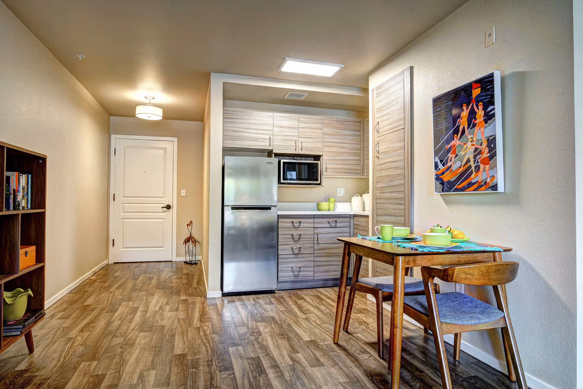 A small kitchen area with light wood cabinets, a stainless steel refrigerator, and a microwave. A wooden dining table with two chairs is set with green cups and bowls. There is a colorful painting on the wall above the table. The floor has a wood pattern, and a white door is visible in the background with a bookshelf on the left side.