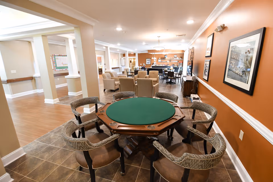 A spacious common area in a senior living facility featuring a green felt octagonal card table surrounded by six cushioned chairs in the foreground. In the background, there are several beige armchairs and dining tables with chairs, a counter area labeled 'Cafe' with shelves and decorations, and warm orange and beige walls adorned with framed artwork.