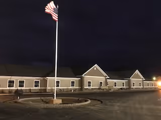 Exterior view of a single-story assisted living facility building at night with an American flag on a flagpole in front and a paved driveway surrounding the flagpole.