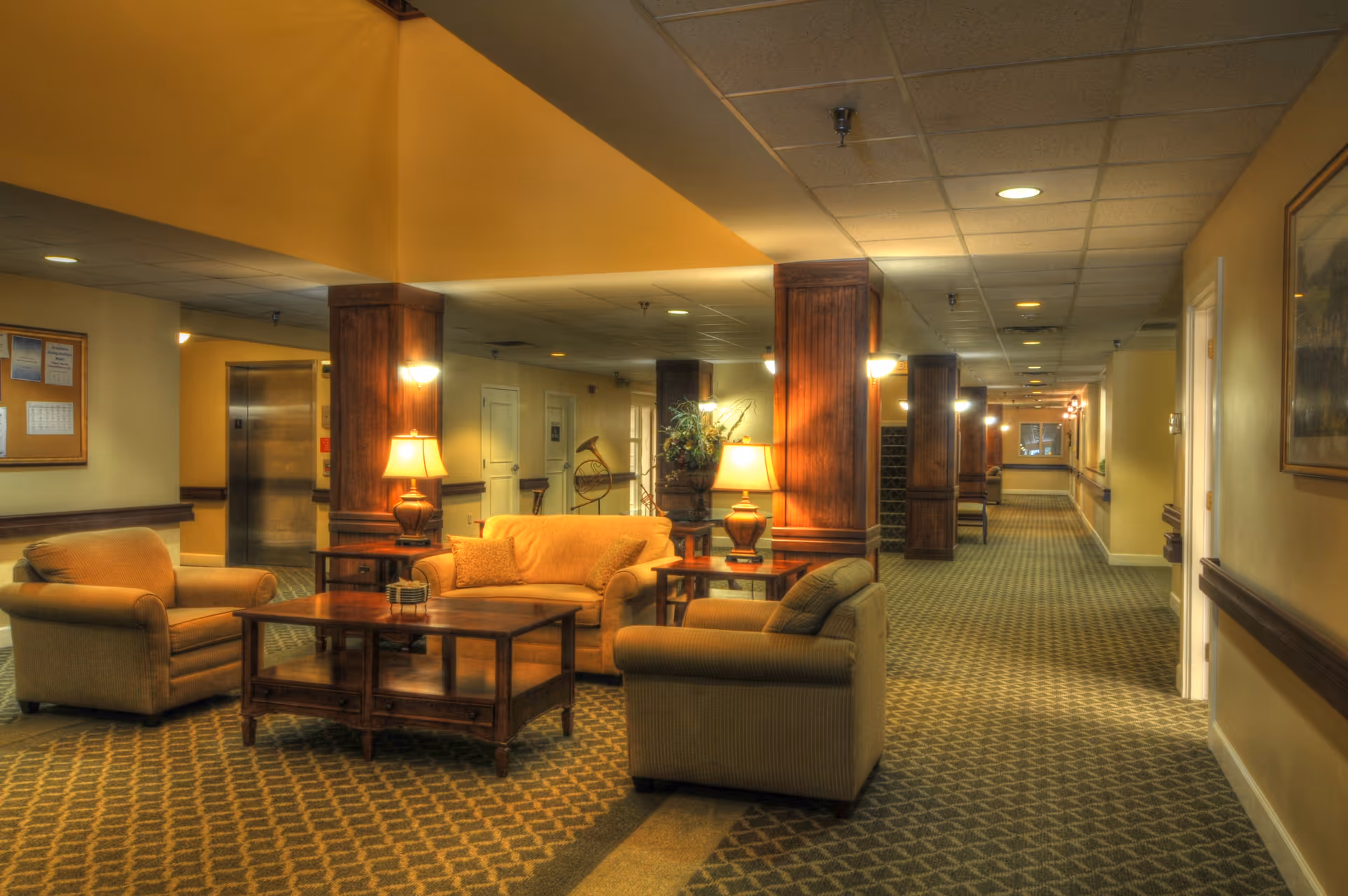 Warmly lit common lounge area in a senior living facility with sofas, armchairs, side tables and lamps along a long carpeted hallway.