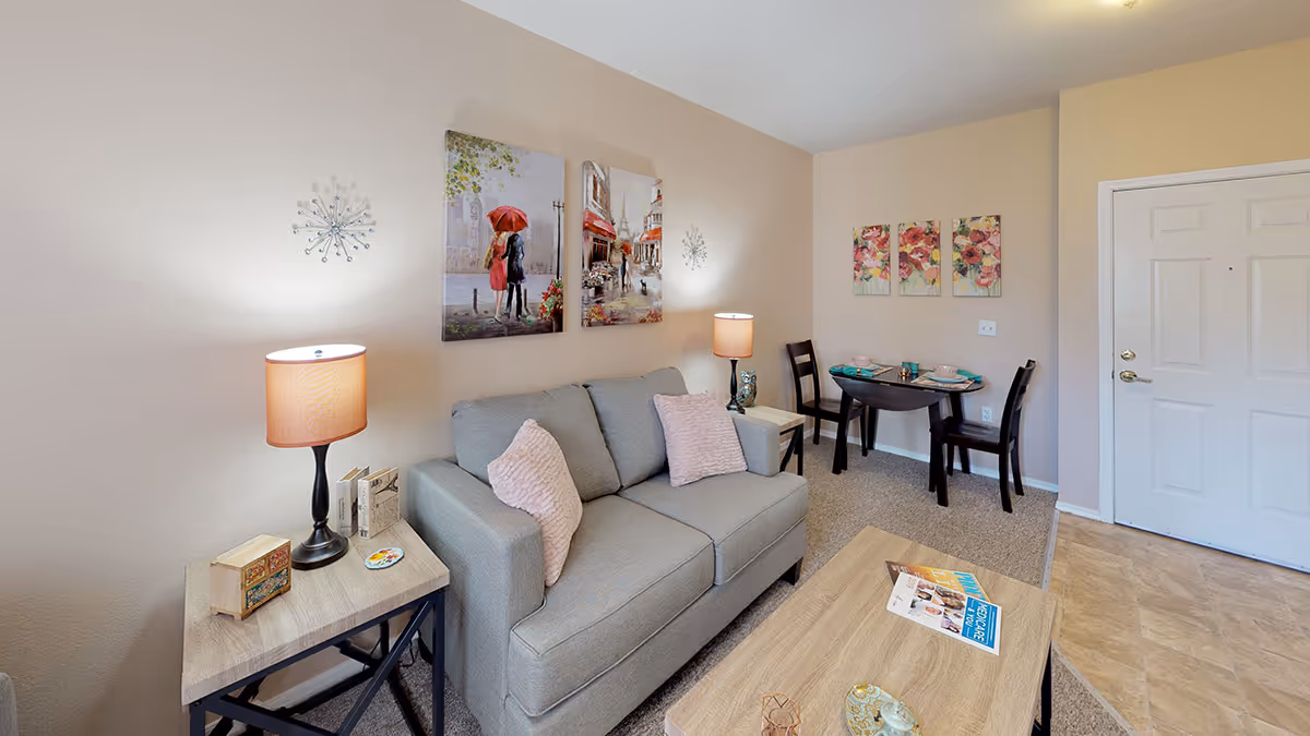 A cozy living room area with a gray couch adorned with two pink pillows, two side tables with lamps, and wall art featuring a couple with an umbrella and a street scene. In the background, there is a small dining table set for two with floral artwork above it and a white door to the right.