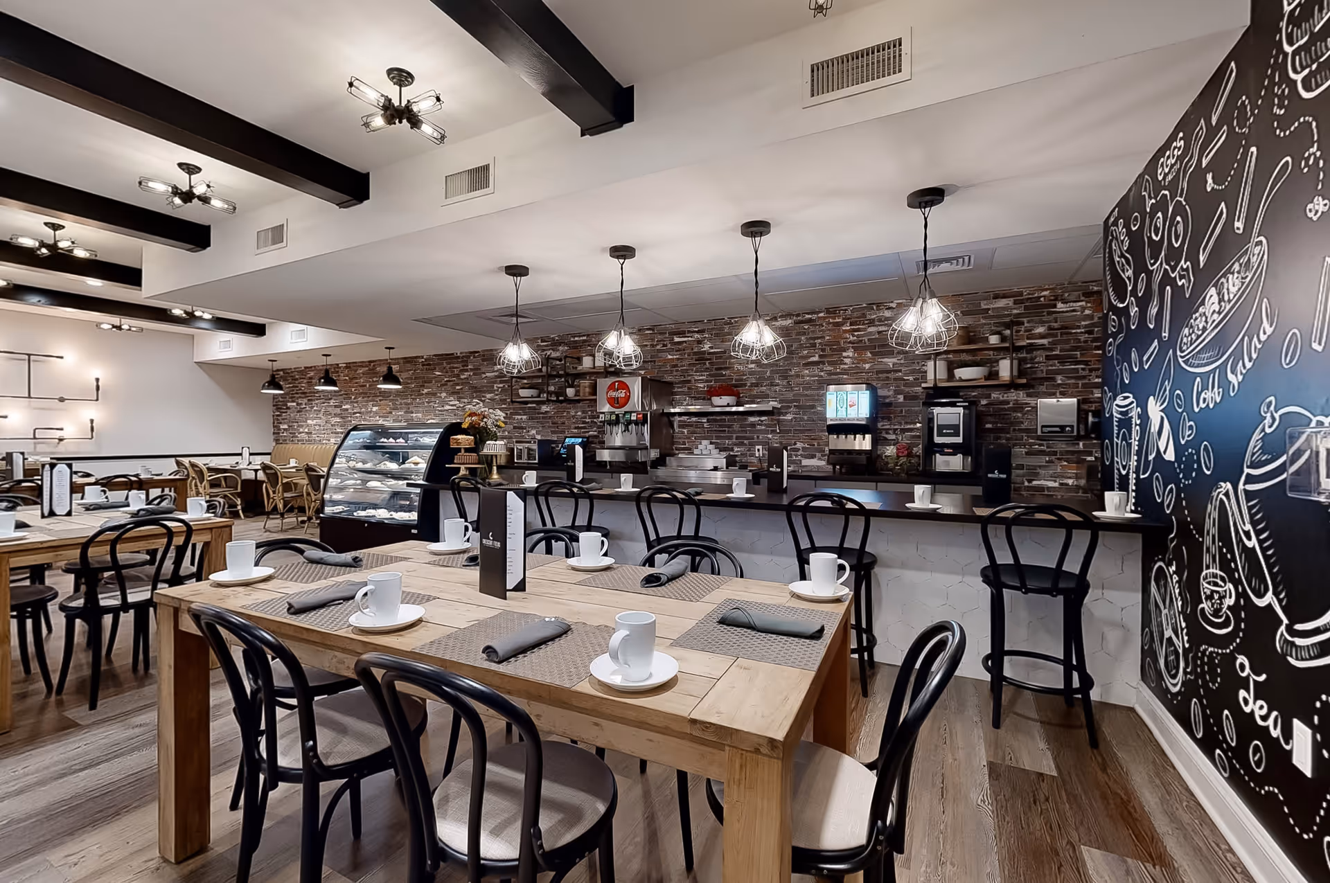 Interior view of a modern dining area with wooden tables set with white cups, saucers, and gray napkins. Black chairs surround the tables. The back wall features a brick design with a counter, beverage dispensers, and a display case with pastries. Pendant lights hang from the ceiling, and a blackboard wall with white chalk-style drawings and text is visible on the right side.