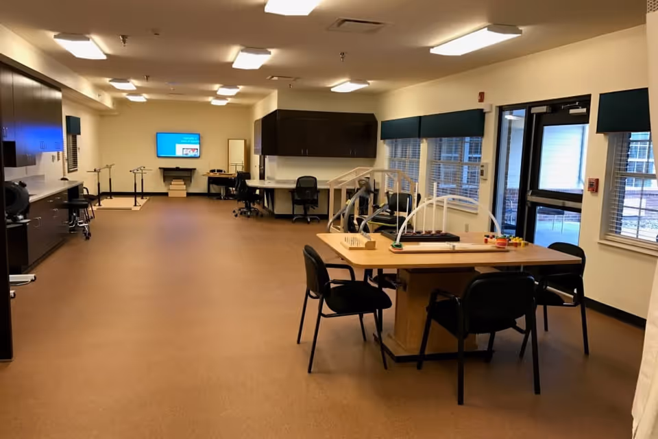 A spacious room with brown flooring and white walls, featuring a wooden table with four black chairs around it. On the table are various activity and therapy tools. The room has large windows with green valances, a door leading outside, multiple desks with chairs, cabinets, and a TV mounted on the far wall. The room appears well-lit with ceiling lights.