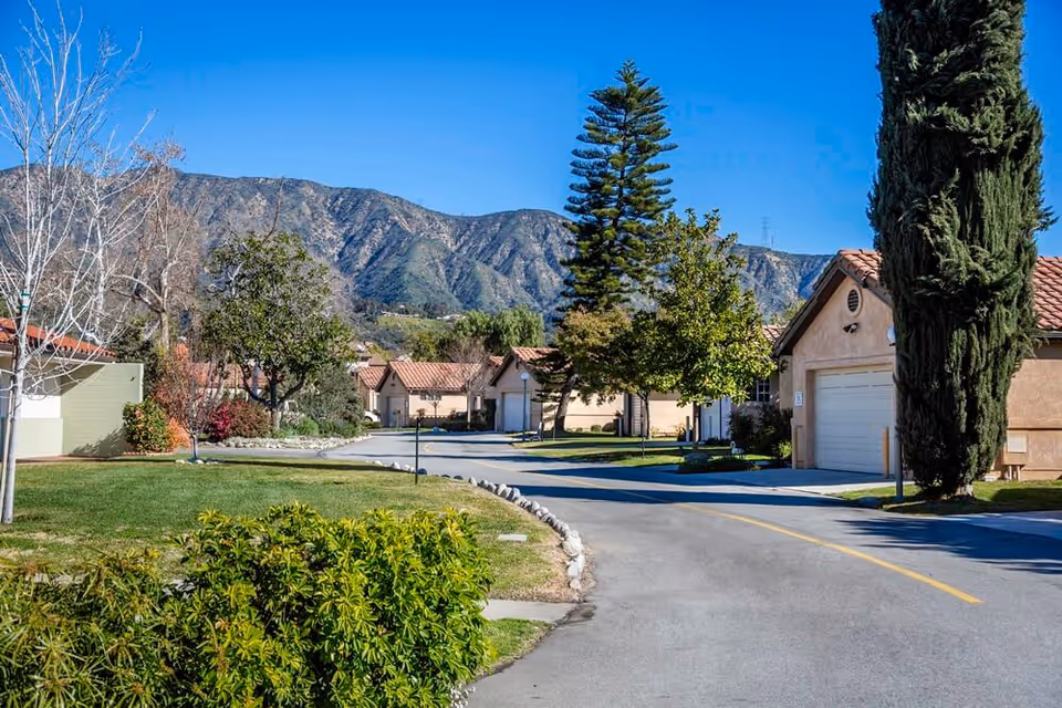 A paved road curves through a residential area with single-story houses featuring tiled roofs. The area is landscaped with green lawns, various trees, and bushes. In the background, there are large mountains under a clear blue sky.