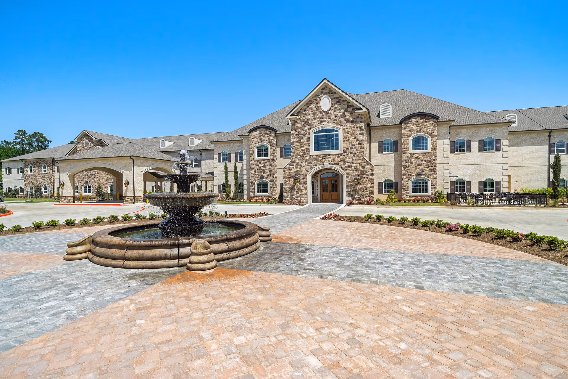 Front exterior view of Worthington Manor, a large two-story building with stone and brick facade, multiple windows, and a central entrance. In front of the building is a circular driveway with a tiered water fountain in the center, surrounded by landscaped bushes and flowers under a clear blue sky.