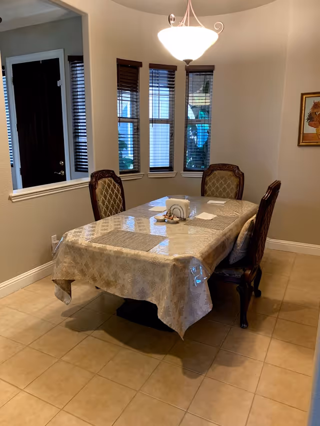 Dining room with a covered rectangular table surrounded by upholstered chairs, bay windows, and a ceiling light.