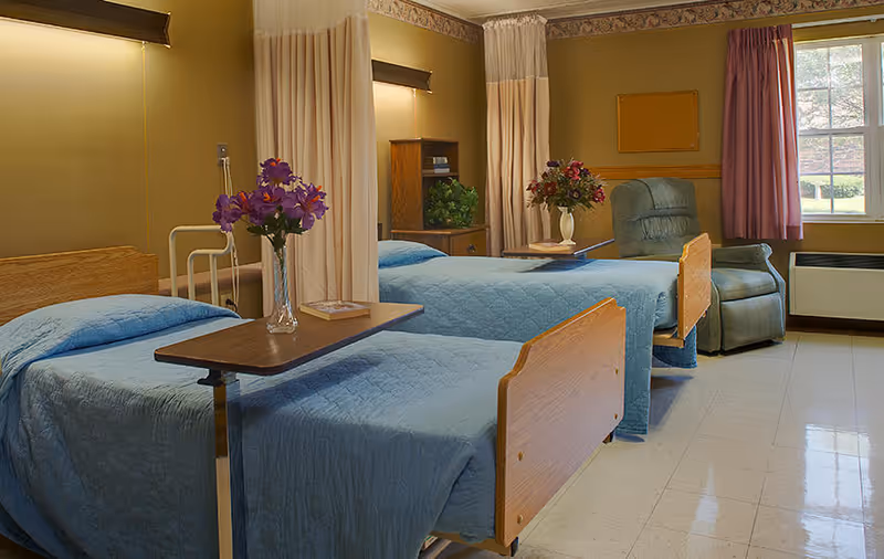 A shared hospital-style room with two beds covered in blue quilts, each with an overbed table holding a vase of flowers. The room has beige walls, a window with purple curtains, a green recliner chair, and a small wooden cabinet with a plant on top. Privacy curtains hang between the beds.