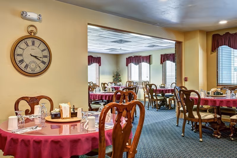 Dining room with round tables covered in burgundy tablecloths, set with glasses, mugs, napkins, and silverware. Wooden chairs surround the tables. The room has windows with burgundy valances, a large wall clock, and a carpeted floor.